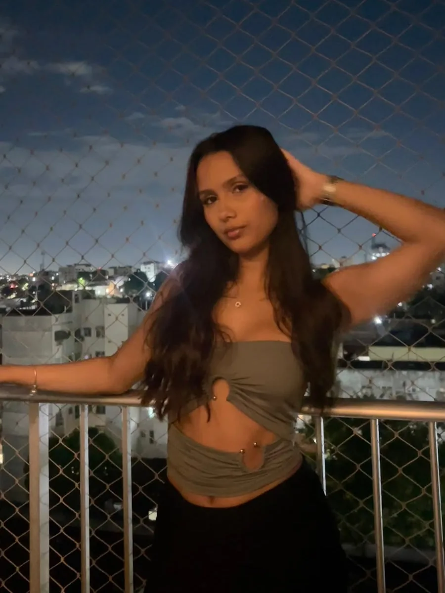 Gabs, streamer da Fusion, de top verde, posando numa varanda à noite com a cidade ao fundo